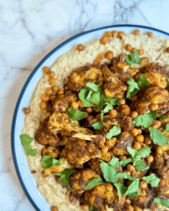 A plate full of homemade chickpea hummus topped with shawarma-spiced cauliflower and fresh herbs.