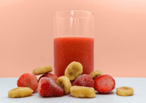 Place UK strawberry and banana smoothie mix, blended in a glass with frozen fruits in front.